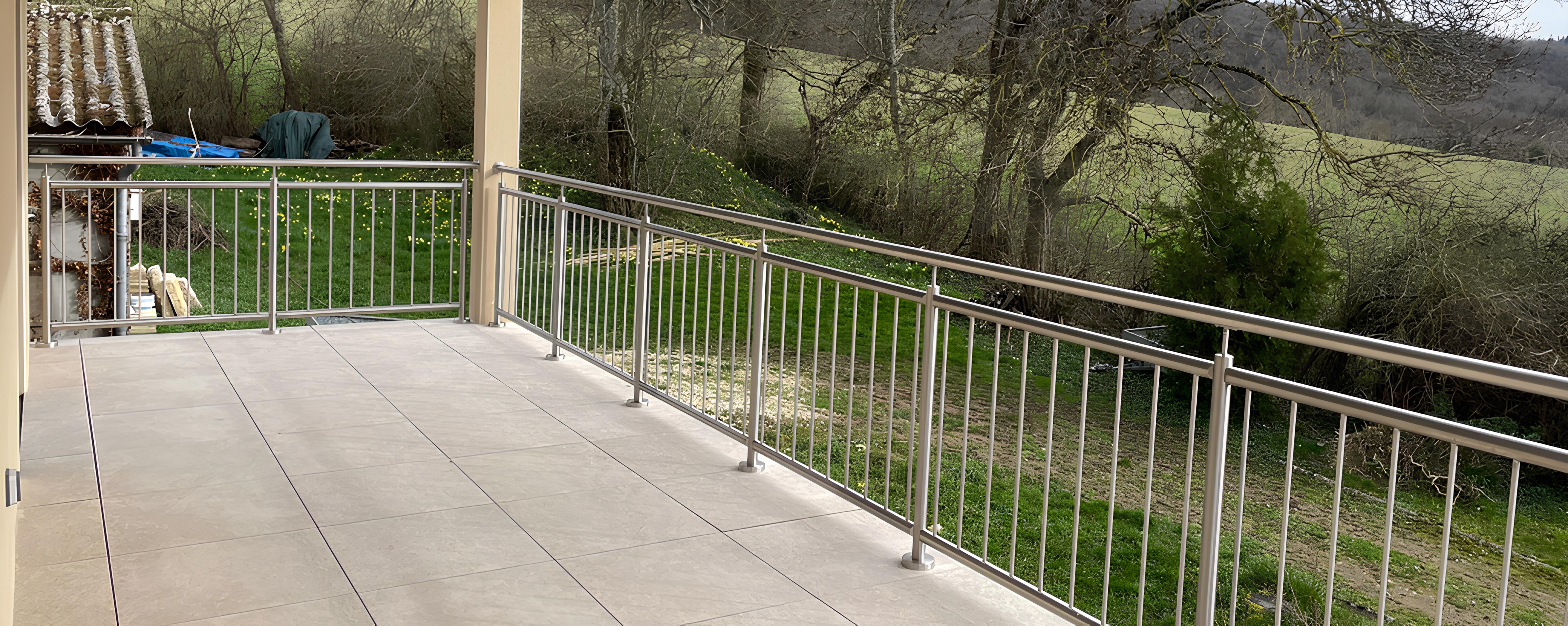 Garde-corps inox à barreaudage vertical sur terrasse carrelée. Garde-corps en inox à barreaudage vertical bordant une terrasse carrelée couverte, avec main courante et poteaux fixés au sol.