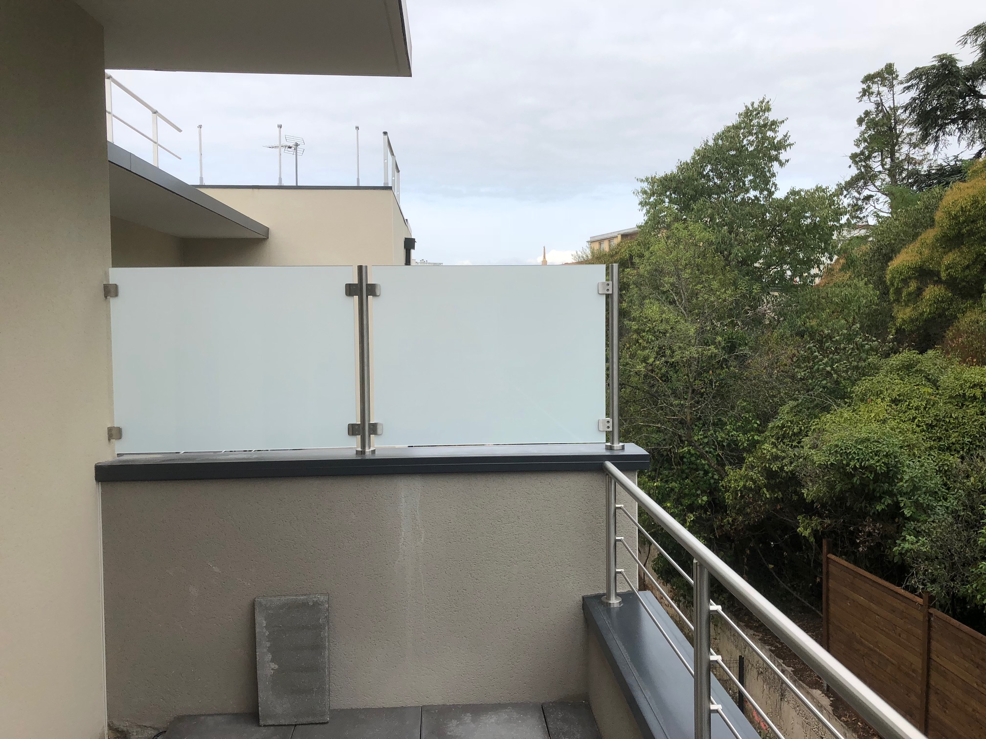 Détail d’un brise-vue en verre 44/2 opale sur acrotère avec couvertine aluminium RAL 7016. Brise-vue en verre opale trempé 44/2 sur acrotère avec couvertine aluminium RAL 7016, avec pinces inox