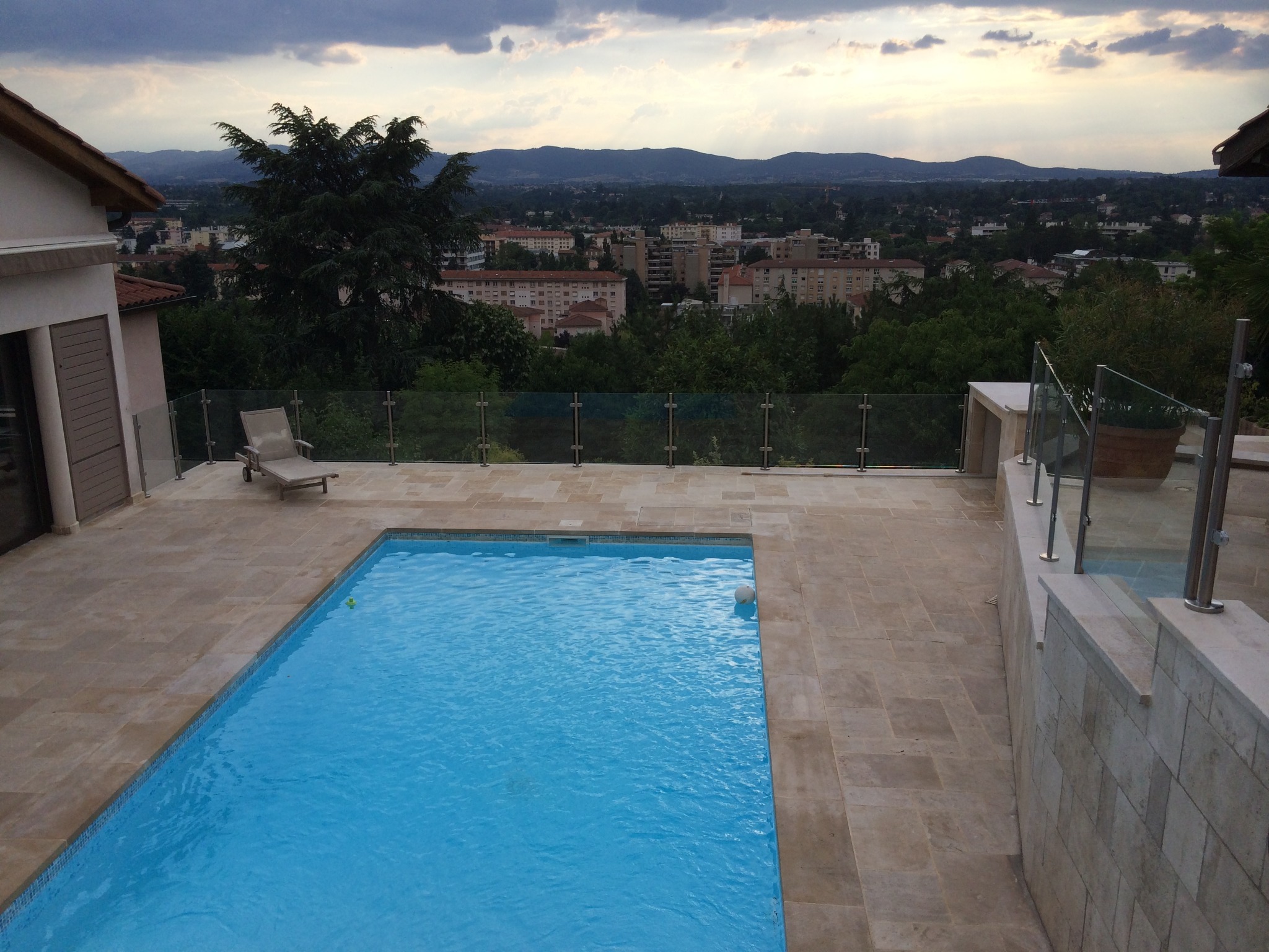 Détail d’un garde-corps tout verre pour piscine sans montants, fixé par pinces à verre. Garde-corps tout verre posé autour d’une piscine, sans montants, sur poteaux inox, pinces à verre, protection piscine aux normes NF P90-306